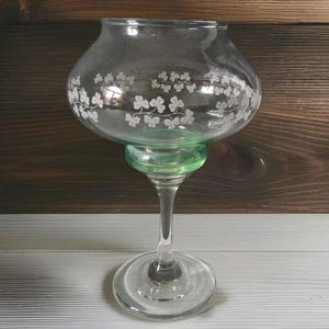May include: A clear glass candle holder with a green base and stem. The bowl-shaped top is decorated with a white clover pattern. The candle holder sits on a round base.
