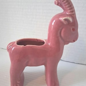 May include: A pink ceramic goat planter with a rectangular opening on its back. The goat has a curved horn, a small tail, and four legs. The planter is a solid, glossy pink color.