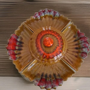 May include: A ceramic serving dish with a flower-like design. The dish is divided into sections, with a central, lidded compartment. The colors include shades of brown, orange, and red. The dish is likely used for serving snacks or appetizers.
