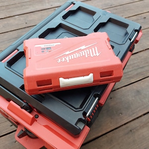 Könnte beinhalten: Ein Stapel roter und schwarzer Milwaukee-Werkzeugkästen. Die obere Werkzeugkiste ist rot mit weißem Griff und dem Milwaukee-Logo. Die unteren Werkzeugkästen sind schwarz und rot. Auf einer Holzoberfläche.