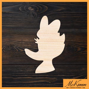 May include: A wooden cutout in the shape of a cartoon bird with a long beak and a curved tail. The bird is made of light-colored wood and is set against a dark brown wood grain background. The logo "McKinnon Design Works" is in the bottom right corner.