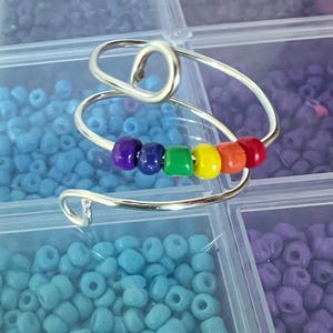 May include: A silver-toned wire ring featuring a spiral design with six small, smooth beads in a rainbow sequence of purple, blue, green, yellow, orange, and red. The ring is constructed from a single continuous piece of metal wire.