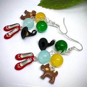 May include: A pair of dangle earrings featuring charms inspired by The Wizard of Oz. The earrings include red shoe charms, a black witch hat, a brown dog, and colorful beads. The earrings are connected by silver-colored chains and hooks.