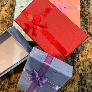 May include: Four small gift boxes with ribbon and bows. The boxes are light blue, red, pink, and a darker blue. The boxes are made of cardboard and have a textured finish.
