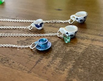 以下が含まれることがあります： ミニチュアのティーカップペンダントが付いたシルバーネックレスが3本。1本のネックレスには、ソーサー付きの青と白のティーカップが付いており、他の2本のネックレスには、青と金のフラワーデザインの白いティーカップが付いています。