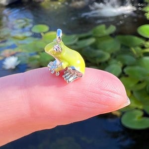 May include: A small, lime green frog pendant with silver-colored accents. The frog charm is perched on a fingertip, set against a blurred background of water and greenery. The pendant has a loop for a necklace.