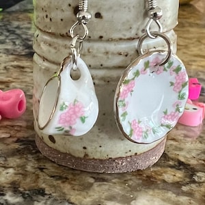 May include: A pair of earrings featuring miniature porcelain teacups with pink floral designs. The earrings are silver and have a hook closure.