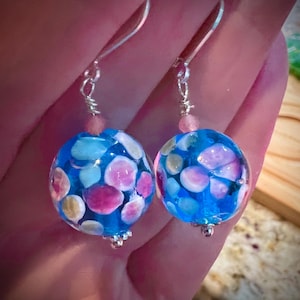 May include: A pair of handmade earrings featuring large, round, blue glass beads. The beads are adorned with colorful, irregular shapes in pink, white, and yellow. The earrings have silver-colored ear wires and small pink beads at the top.