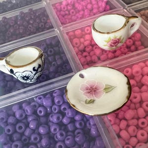 May include: Miniature porcelain teacups and a small plate with floral designs. The teacups have gold trim and painted flowers. The plate has a pink flower design. The items are displayed in a divided container with colorful beads.