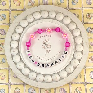 May include: Pink and white beaded bracelet with the words "I AM DRAMATIC" and the text "Swiftie Bracelets Co."