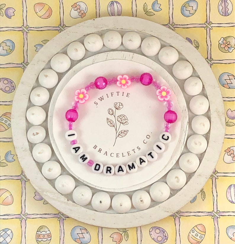 May include: Pink and white beaded bracelet with the words "I AM DRAMATIC" and the text "Swiftie Bracelets Co."