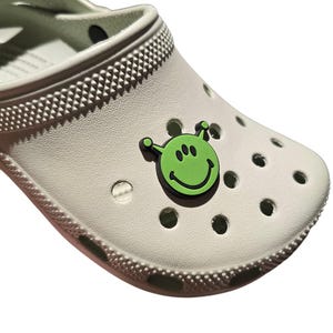 May include: A white clog with a green alien smiley face charm. The charm has black outlines and three black eyes.