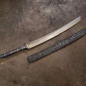 Puede incluir: Una espada katana artesanal con una hoja plateada y un mango oscuro con dibujos. La espada va acompañada de una vaina negra a juego con una textura tallada. La hoja mide aproximadamente 71 cm de largo.