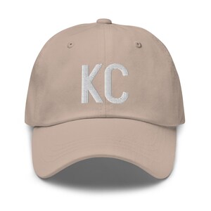 May include: A beige baseball cap with white embroidered letters spelling "KC".