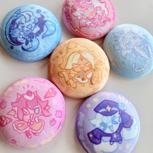 May include: A collection of seven plush, round pins featuring cartoon character designs in various colors. The pins have a soft, textured appearance, with designs in shades of blue, pink, orange, and purple. The pins are arranged on a white surface.