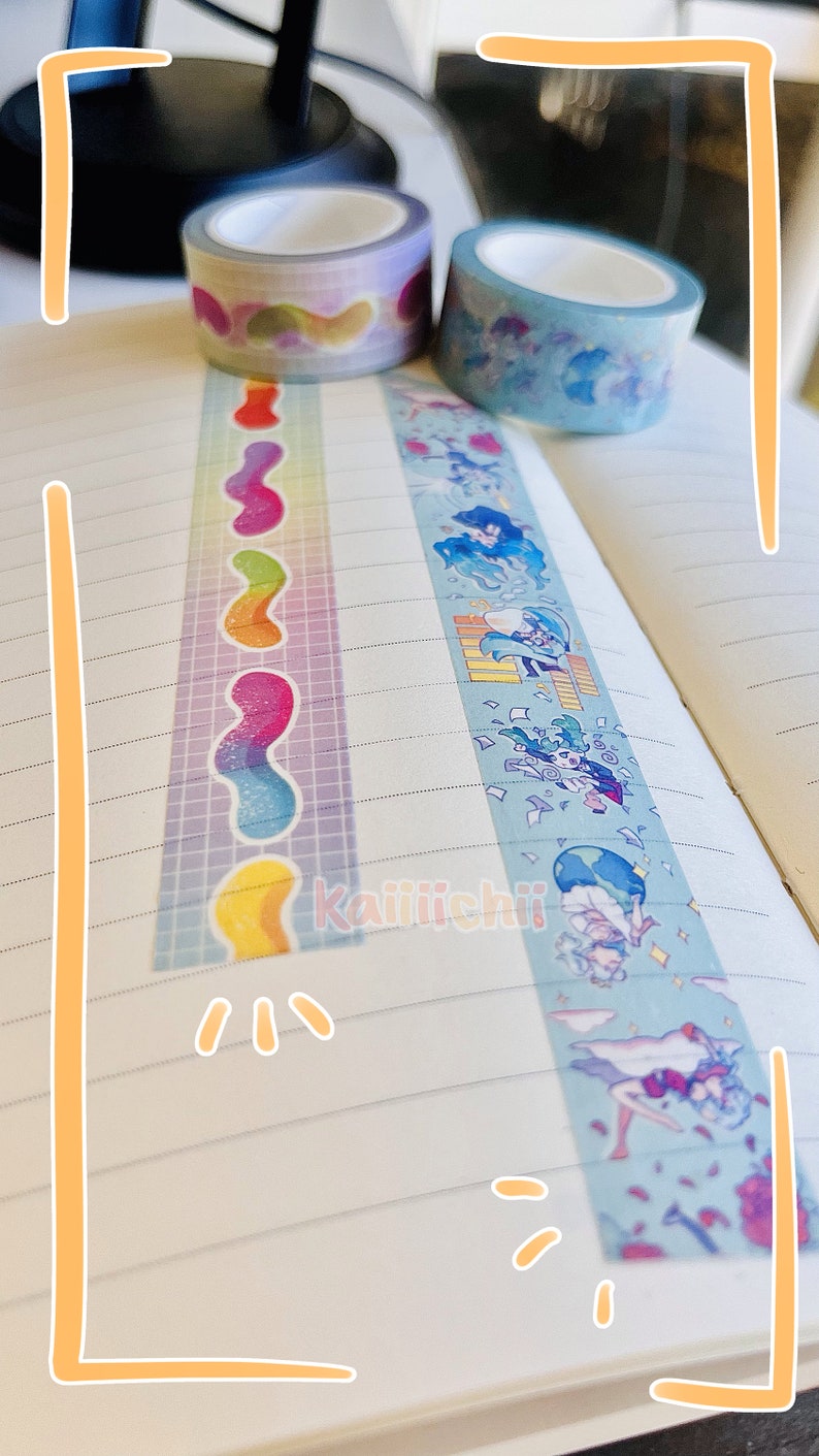 May include: Two rolls of decorative washi tape. One roll has a rainbow pattern with a wavy design. The other roll has a blue background with a cartoon character design. The text "Kaiiichii" is visible on the page.