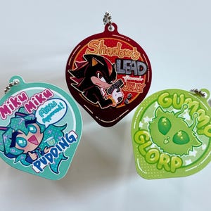 May include: Three colorful keychains with cartoon graphics. One is teal with a character and the words "Miku Pudding". Another is maroon with "Shadow's Lead Almond Jelly". The last is green with "Gummy Glorp".