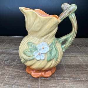 Puede incluir: Jarra de cerámica con diseño floral. La jarra es principalmente amarilla con detalles en naranja y verde. Tiene un asa y un pico, y está decorada con flores blancas y hojas verdes. La base es naranja.