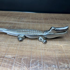 May include: A silver-colored alligator figurine with detailed scales and open jaws. The figurine is lying on a wooden surface, showcasing its realistic design and metallic finish. The alligator has four legs and a long tail.