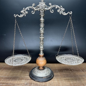 May include: A decorative balance scale featuring a silver-toned metal frame with ornate scrollwork at the top, a central wooden sphere accent, and a dark circular base. Two clear, patterned glass plates are suspended by thin metal chains on each side.