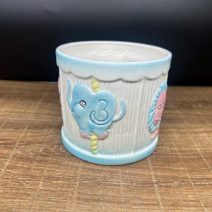 Könnte beinhalten: Keramik-Pflanztopf in Hellblau und Weiß. Er hat geprägte Karusselltier-Motive, darunter einen blauen Elefanten und einen rosa Löwen. Der Rand und die Basis sind hellblau, der Körper hat vertikale Linien.