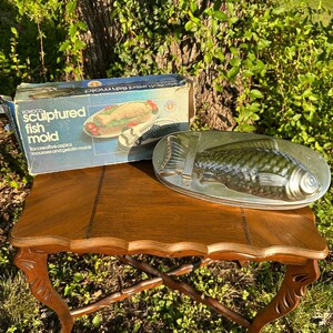 May include: A vintage fish mold set, including a silver-colored fish-shaped mold and a box with the text "Sculptured Fish Mold". The set is displayed on a wooden table with ornate legs, set outdoors.