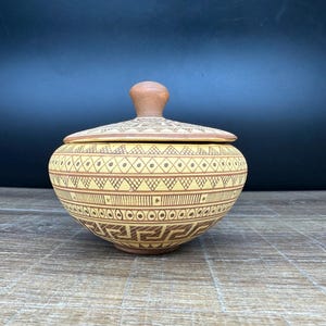 May include: A tan ceramic bowl with a lid. The bowl and lid are decorated with brown geometric patterns. The lid has a small, rounded handle. The bowl is sitting on a wooden surface.