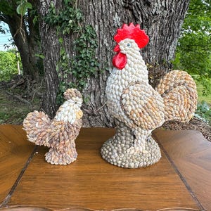 May include: Two rooster sculptures crafted from seashells. The larger one features a red comb and wattle, with a body of white and tan shells. The smaller rooster is brown and white. Both are on a wooden table.