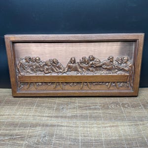 May include: A rectangular wall plaque featuring a relief carving of The Last Supper. The scene depicts thirteen figures seated at a long table, rendered in a monochromatic brown tone. The artwork is encased in a simple, matching brown wooden frame.