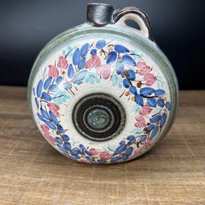 Op de afbeelding: Een ronde keramische kruik met een handvat en een kleine tuit. De kruik is versierd met blauwe en roze bloempatronen op een crèmekleurige achtergrond. De rand en het handvat zijn bruin.