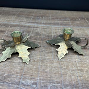 Puede incluir: Un par de candelabros de latón con forma de hojas de acebo. Cada candelabro cuenta con una copa cilíndrica central para una vela cónica, una base plana diseñada con venas de hoja detalladas y bordes dentados, y una pequeña asa circular lateral.