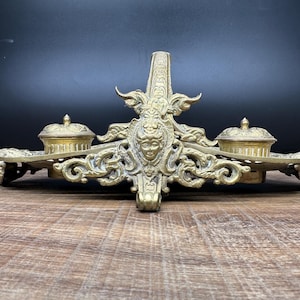May include: A gold-toned metal desk inkwell stand featuring an ornate central relief of a human face surrounded by intricate scrollwork. The piece includes two lidded circular ink containers and a central vertical pen holder on a wide base.