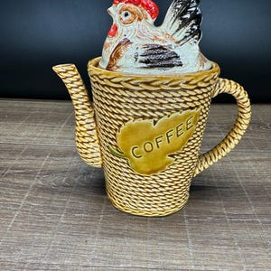 May include: A ceramic coffee pot with a textured, woven-look exterior in a tan finish. The pot features a prominent leaf-shaped emblem with the word COFFEE embossed on the front. A lid shaped like a chicken with a red comb and black tail sits on top.