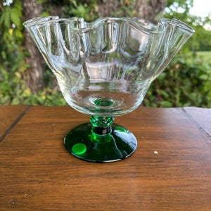 May include: A clear glass dessert bowl with a ruffled rim, set on a green stem and base. The bowl is transparent, allowing light to pass through. The base is a solid, dark green color.