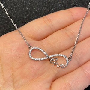 Könnte beinhalten: Eine silberfarbene Halskette mit einem Unendlichkeitssymbol, das mit kleinen, klaren Kristallen besetzt ist. Das Wort "love" ist in Schreibschrift innerhalb der Unendlichkeitsschleife geschrieben. Die Halskette hat eine feine Kette.