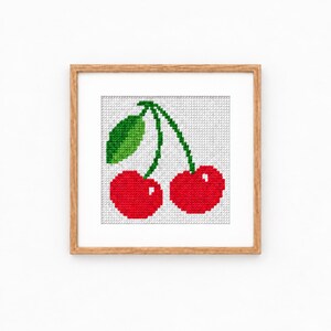 Puede incluir: Un marco de madera cuadrado que contiene un bordado de punto de cruz de dos cerezas de color rojo brillante con tallos verdes y una hoja verde. Las cerezas están representadas en un estilo pixelado sobre un fondo de cuadrícula blanca.