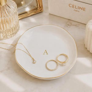 May include: A round white ceramic trinket dish with a gold-trimmed edge and the letter A printed in gold in the center. The dish holds a delicate gold chain necklace with a small clear stone pendant and two gold rings, one plain and one with a stone.