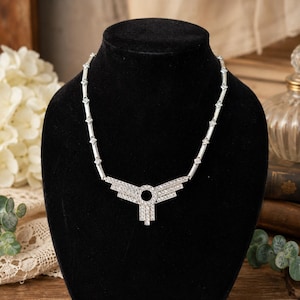 May include: A silver-toned necklace featuring a central geometric pendant with a circular cutout and tiered, crystal-encrusted bars. The chain consists of alternating smooth silver-toned segments and small, square-shaped crystal accents.