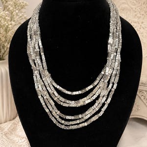 May include: A multi-strand silver-toned necklace displayed on a black mannequin. The necklace features multiple rows of rectangular, clear beads. The beads are arranged in a cascading design, creating a layered effect. The background includes white flowers and a decorative white dish.