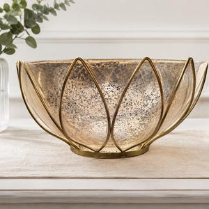 May include: A decorative glass bowl with a gold-colored metal frame. The bowl has a textured, speckled appearance and a wide, open design. The metal frame forms a petal-like shape around the glass. The bowl is designed for decorative purposes.