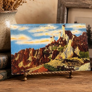 May include: Vintage postcard featuring a colorful landscape of rock formations under a blue sky with clouds. The postcard is displayed on a decorative brass stand. The text on the postcard reads "HELL'S HALF ACRE IN WONDERFUL WYOMING."