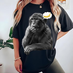 May include: Black t-shirt featuring a grayscale image of a monkey in a thinking pose, with a thought bubble showing a yellow banana. The monkey is holding its chin, suggesting contemplation. The shirt is a casual style.