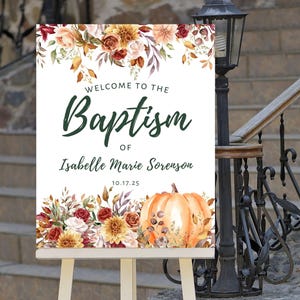 Op de afbeelding: Een wit bord met herfstachtige bloemenversiering en een pompoen. Het bord staat er: "Welkom bij de doop van Isabelle Marie Sorenson 10.17.25".