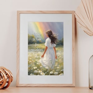 Op de afbeelding: Een ingelijst afdruk met een aquarel van een vrouw die door een veld met madeliefjes loopt. Achter haar is een regenboog te zien in de lucht. De tekst "Ik hou ervan om naar regenbogen te zoeken" staat in wit op de afdruk.