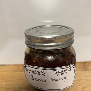 May include: A glass jar with a silver lid filled with a dark brown sauce. The jar has a white label with the text "Saucey Jawns Jeow bong" written in black.