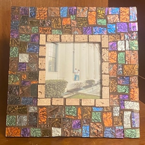 Puede incluir: Marco de fotos cuadrado de mosaico con una foto de una pareja. El marco está hecho de pequeñas baldosas de colores en tonos marrones, naranjas, morados y verdes. El marco interior es de color marrón claro.