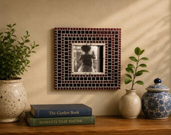 May include: A square picture frame featuring a mosaic border of small red and black tiles surrounding a central white border. The frame contains a black and white photograph of a child. The frame is mounted on a wall above two stacked books.
