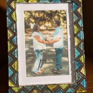 May include: A rectangular photo frame with a mosaic border. The frame's border features a zigzag pattern of blue, green, and brown glass tiles. The frame holds a photograph of a couple holding hands. The frame is displayed on a wooden surface.