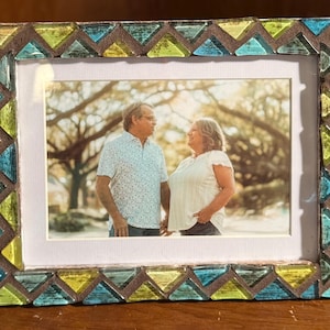 May include: A rectangular picture frame featuring a mosaic border composed of alternating triangular glass tiles in shades of lime green and turquoise blue, set against a dark brown base, surrounding a photograph of two people standing outdoors.