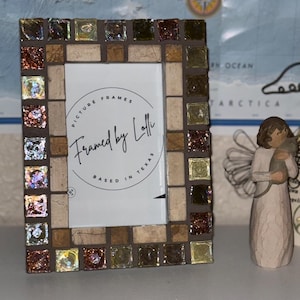 Puede incluir: Un marco de fotos rectangular con un borde de mosaico de azulejos de vidrio iridiscente cuadrados en tonos marrón, dorado y cobre, intercalados con azulejos de piedra beige. El inserto central muestra el texto Framed by Lolli Based in Texas.
